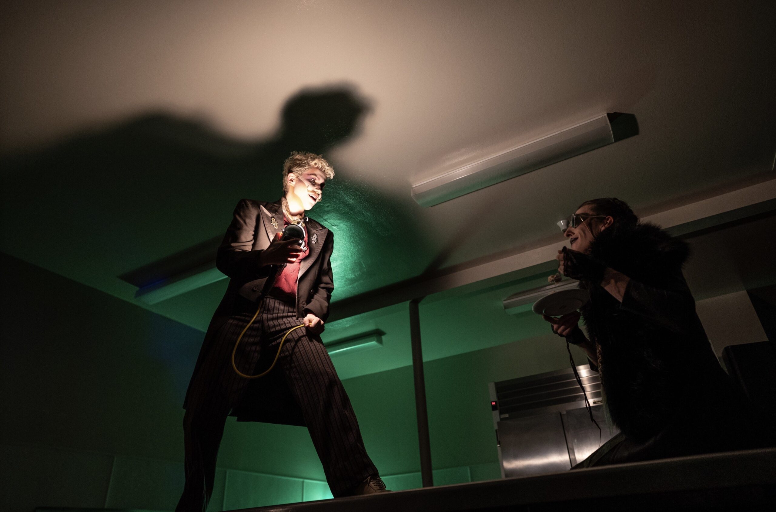 In this dramatic scene, Syd Franz stands on a table holding a lightbulb with a long cord, casting a large shadow on the ceiling as they face another person (Quinn Dixon) in dark clothing and sunglasses, who appears to be reacting with intensity, adding to the suspenseful atmosphere.